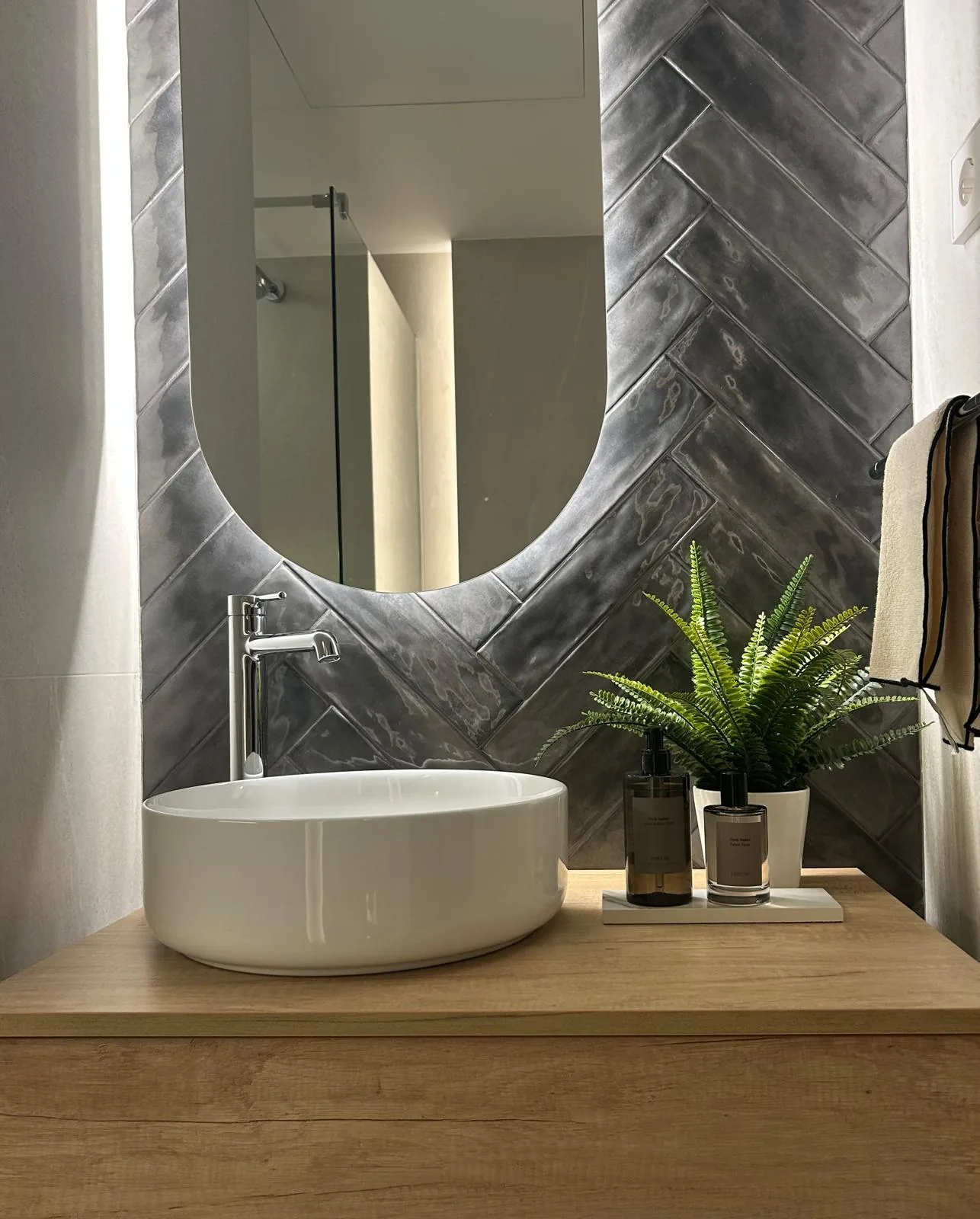 Baño con espejo ovalado, revestimiento gris en espiga, lavabo de sobre y mueble de madera