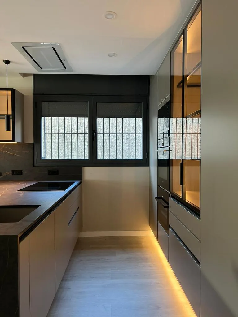 Cocina con encimera oscura, horno en columna, ventana con reja y luz led en el suelo